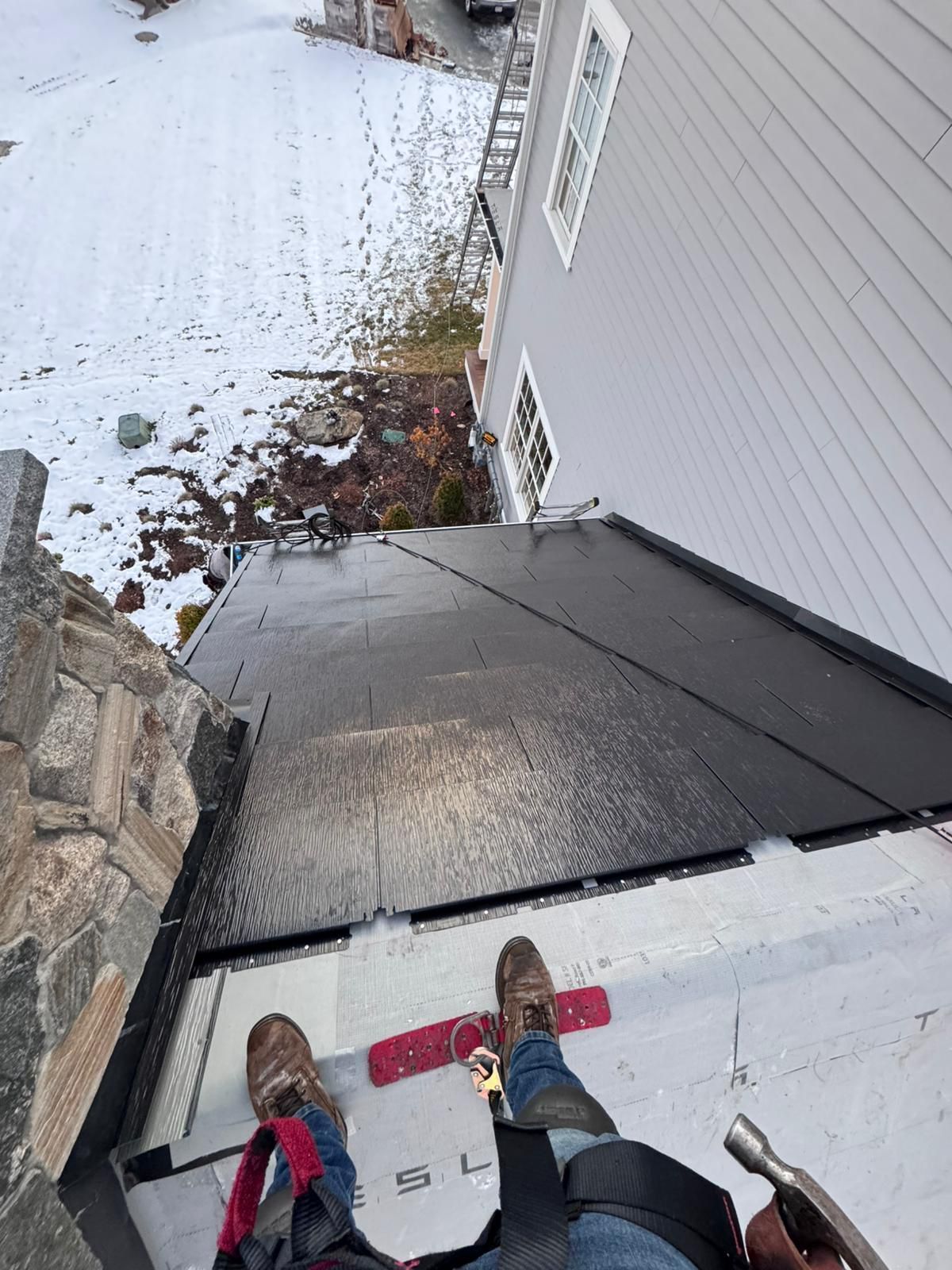 Person on a roof wearing a safety harness, hammer in hand, near a snowy landscape.