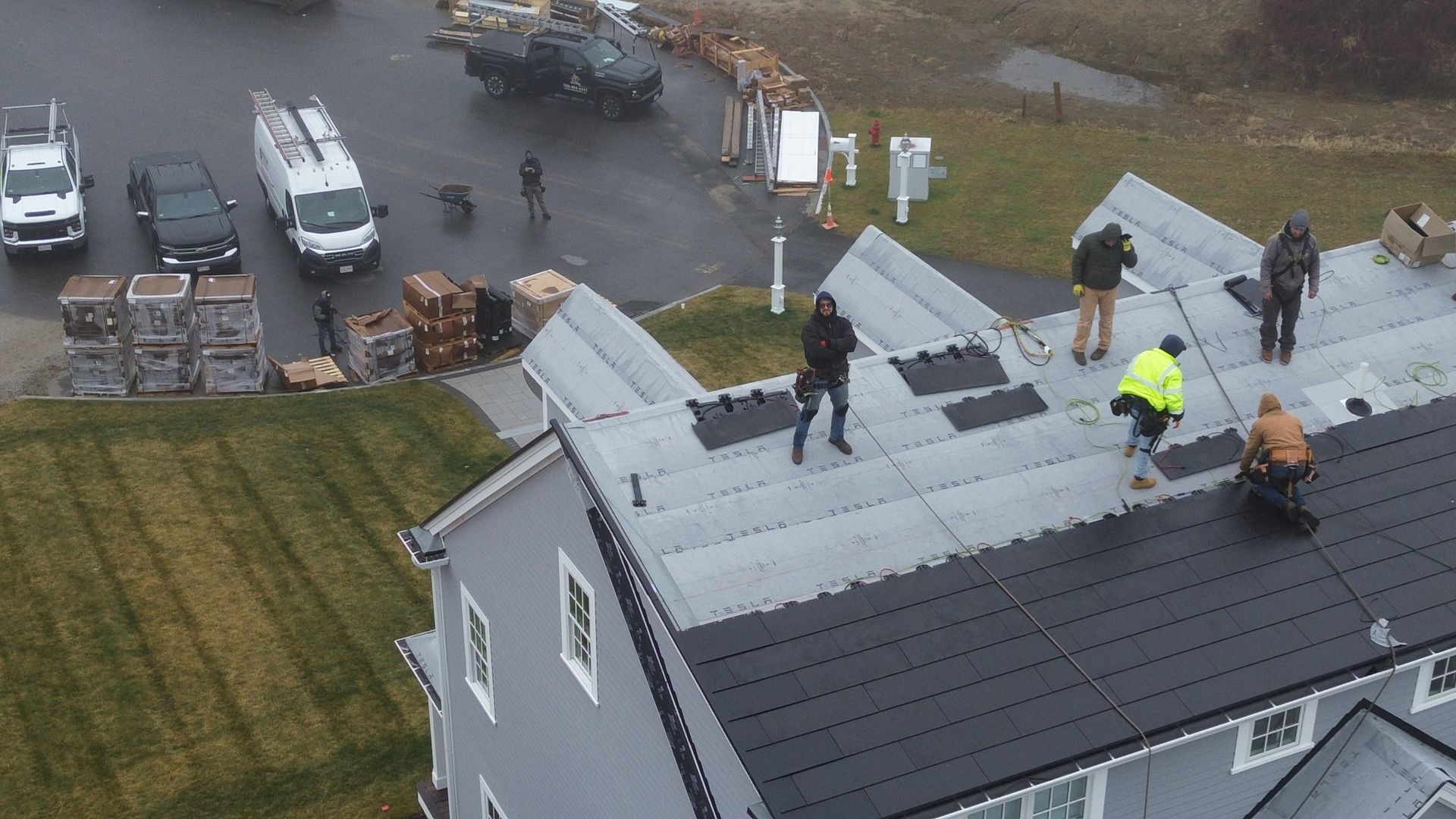 Solar panel installation on a gray roof; workers in jackets. Trucks and equipment parked nearby.