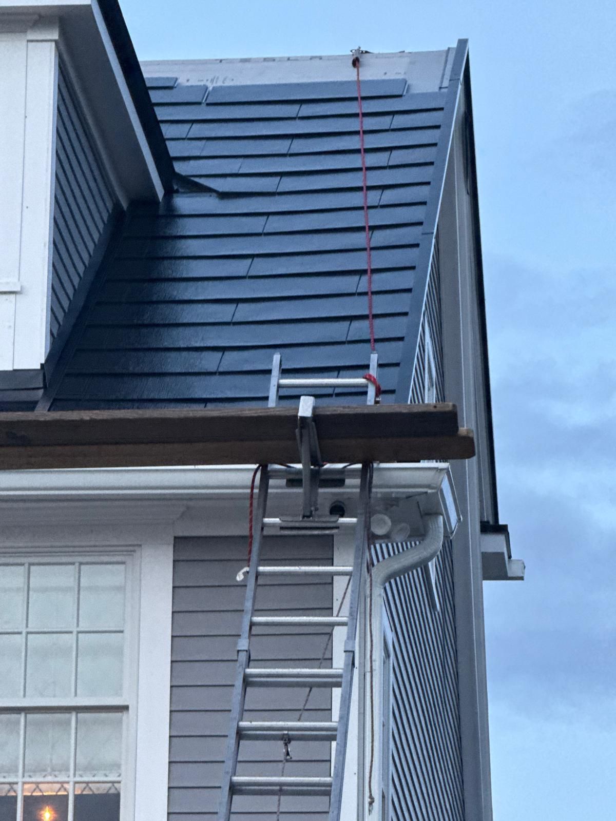 Ladder and plank set up on house roof for construction. Red rope secured to roof. Grey siding.