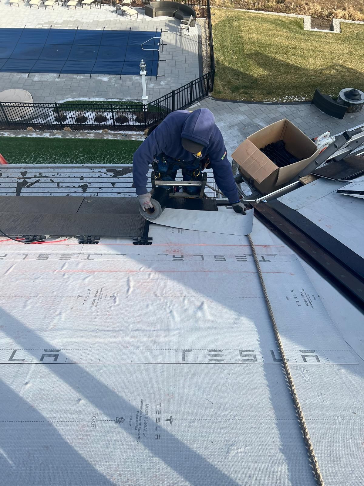 Person in blue working on a roof, cutting material with a tool. Box and pool in the background.