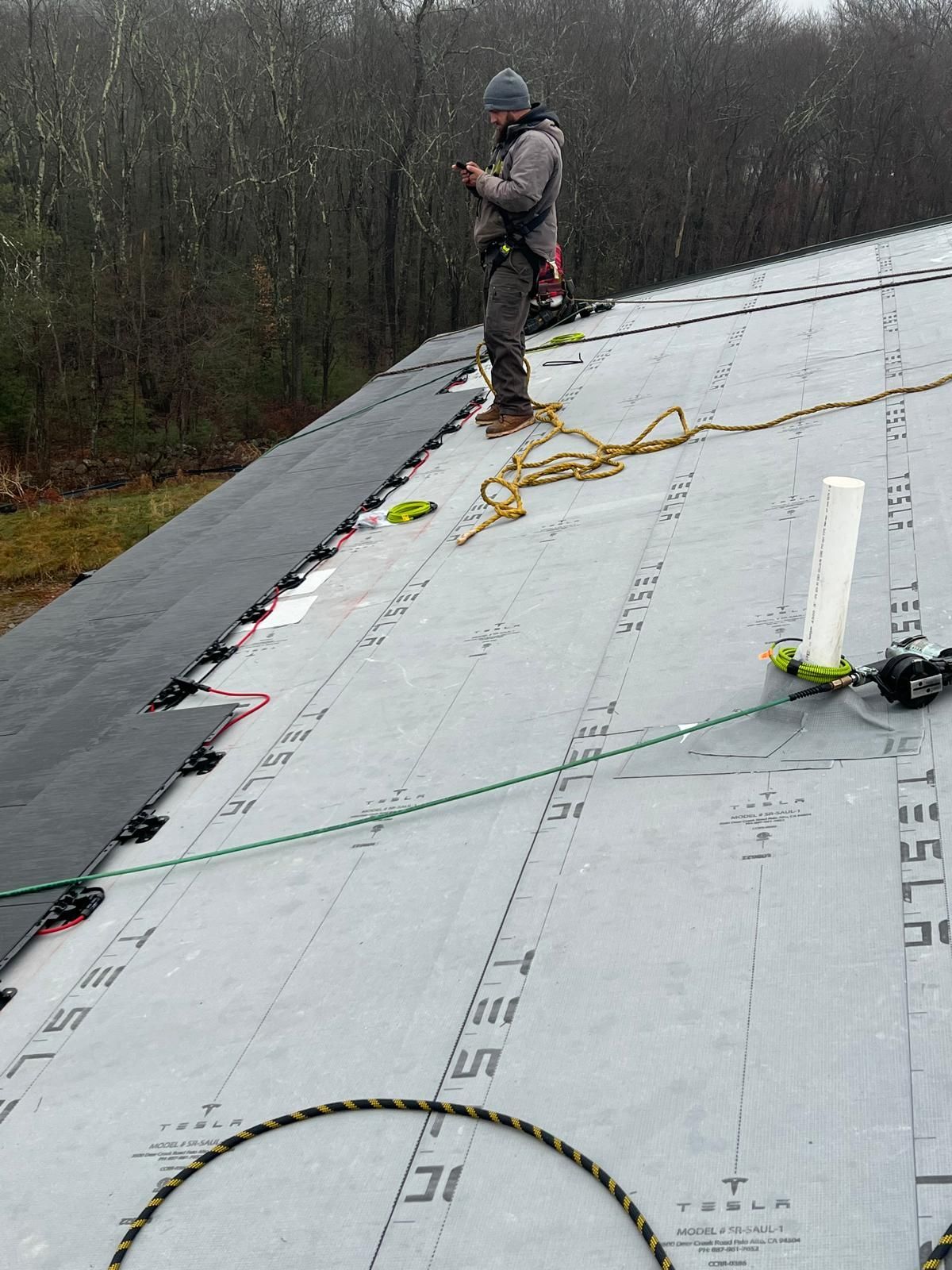 Person on a roof installing solar panels. Harness, safety rope, tools. Forest background, overcast day.