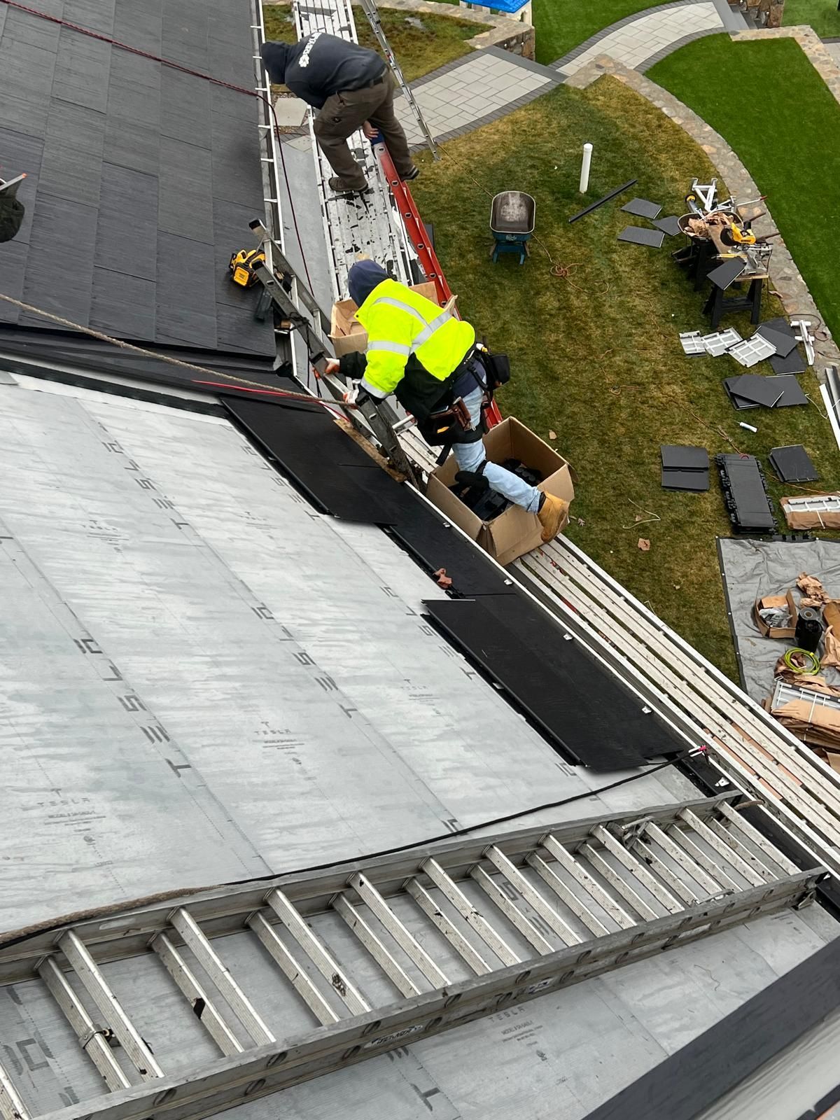 Two roofers on a sloped roof. One in yellow vest handles materials, other at top. Silver ladder present.