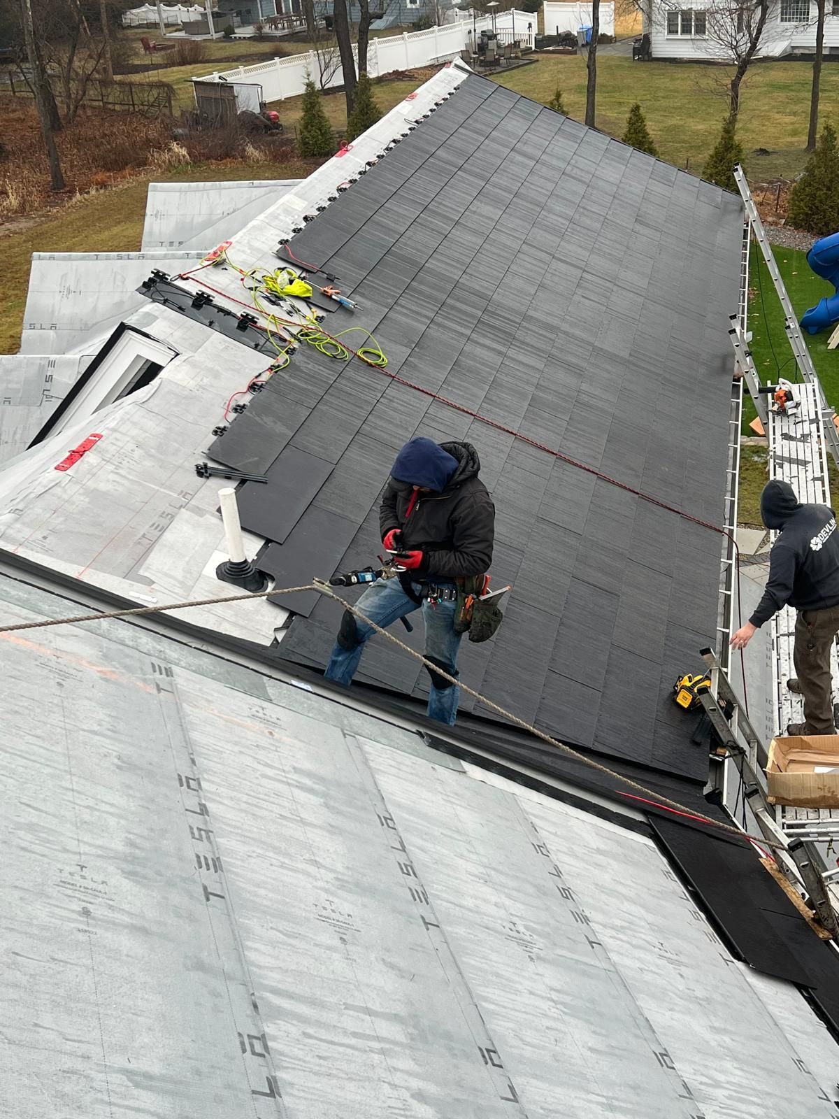 Roofer on a sloped roof wearing safety gear, working on roofing shingles. Outdoors, daylight.