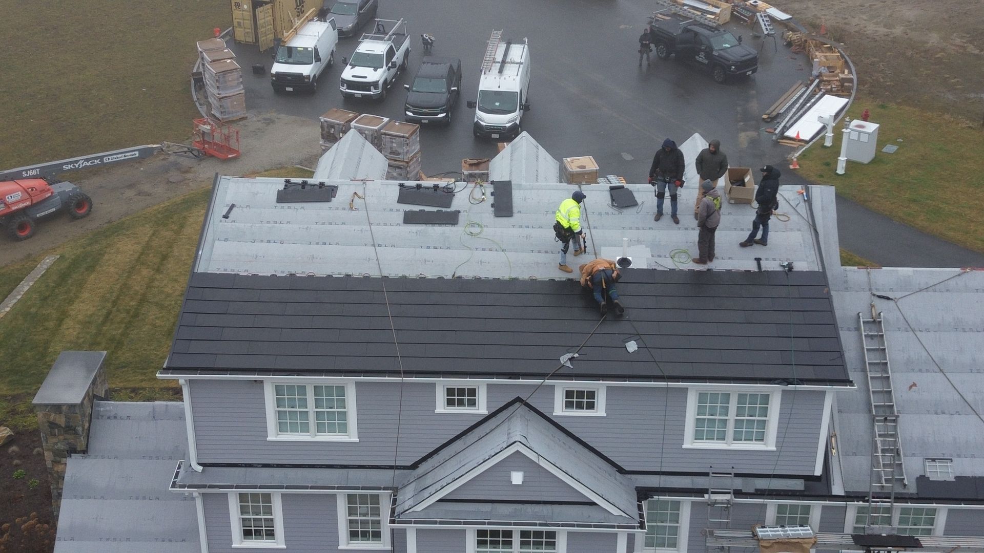 Roofers installing black shingles on a gray house roof. Vehicles and equipment are in the background.