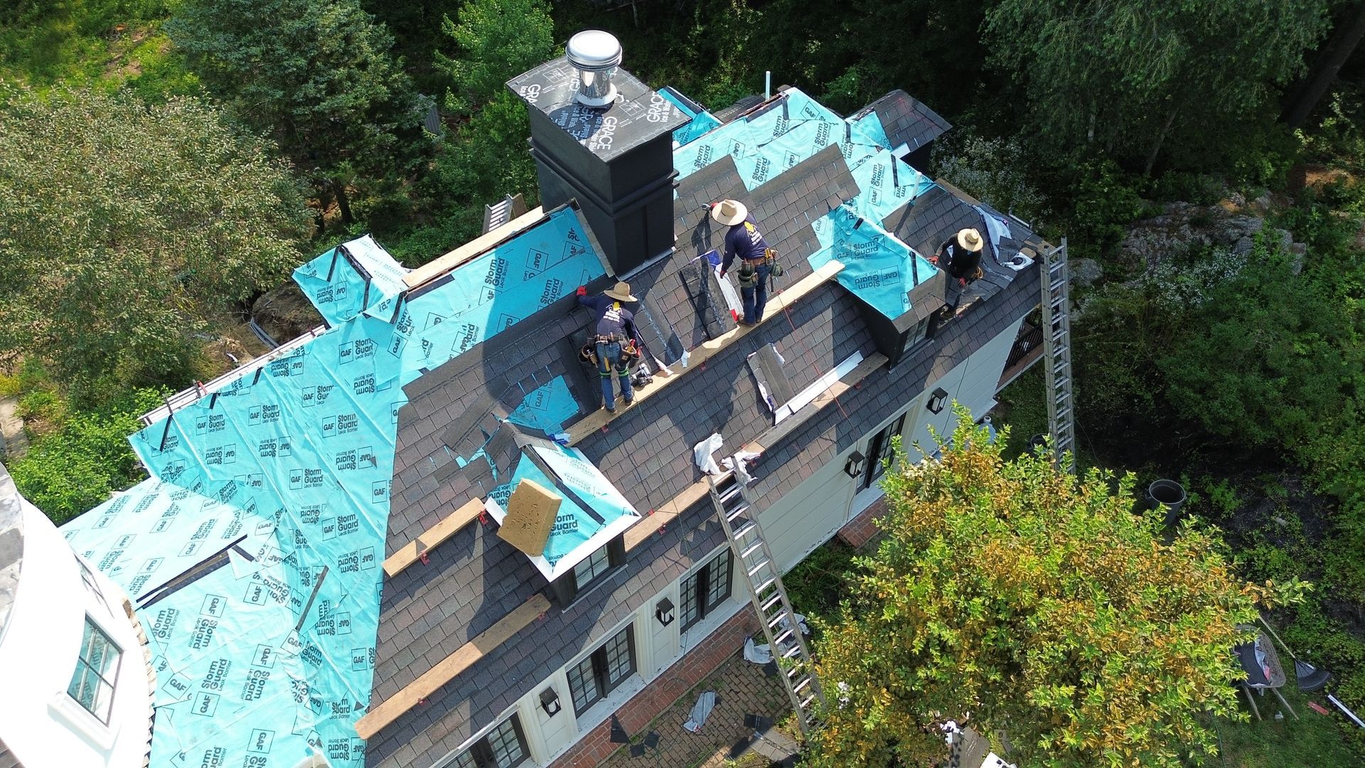 Roofers installing shingles on a house roof, surrounded by green trees.