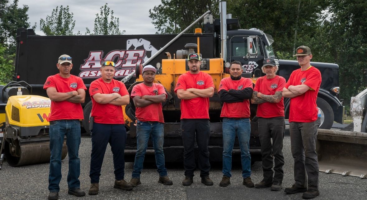 Paved driveway leads to a gray house, surrounded by trees and green grass, under a bright blue sky.