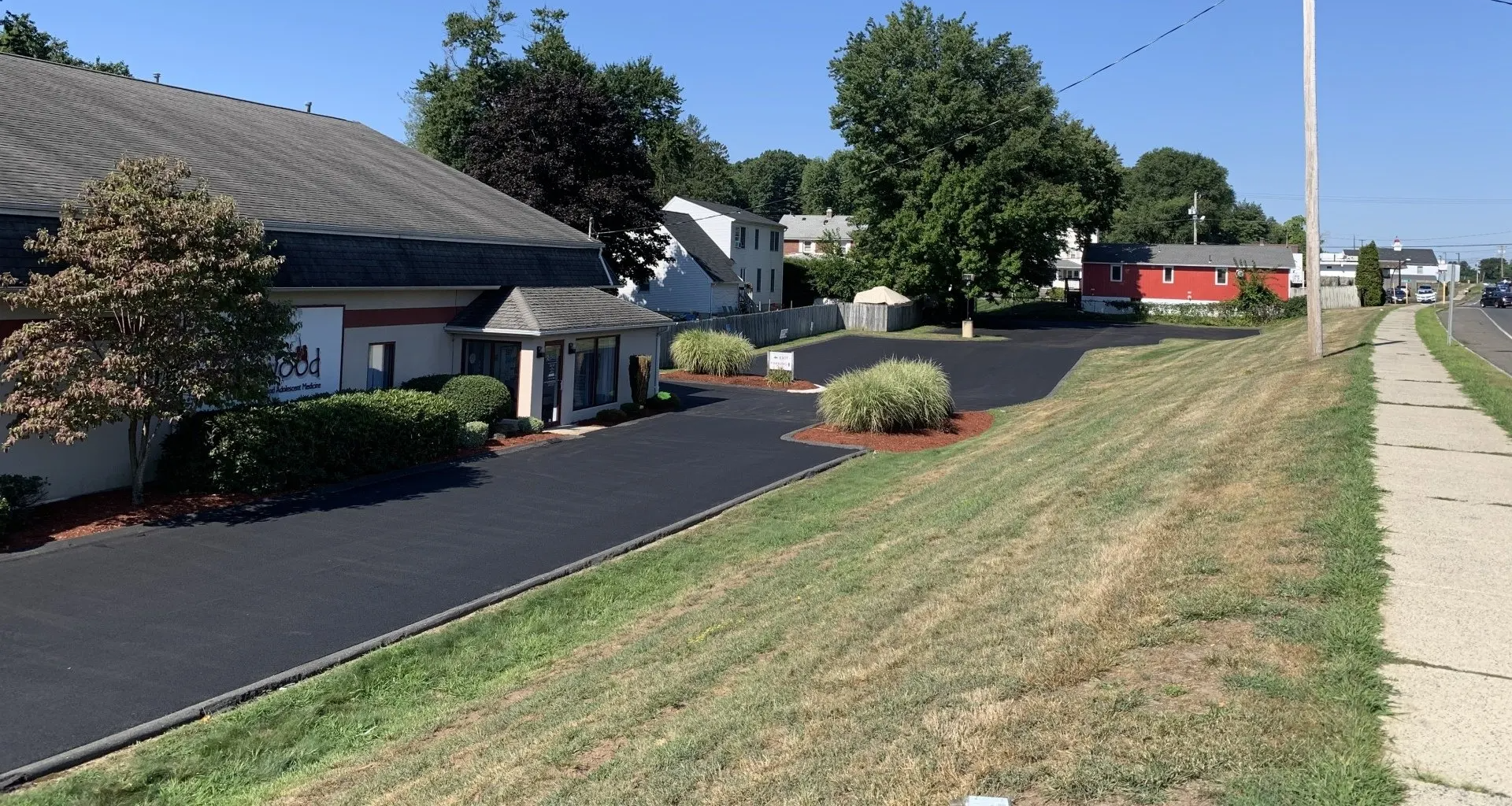 Asphalt driveway leads to low building, then to homes on a sunny day.  Grassy verge and sidewalk on right.