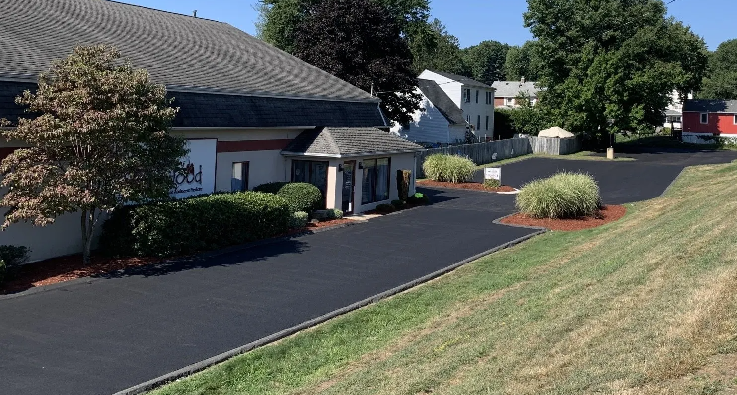 A building with a dark roof and entrance under a canopy; a driveway extends into the distance, with grassy areas and trees.