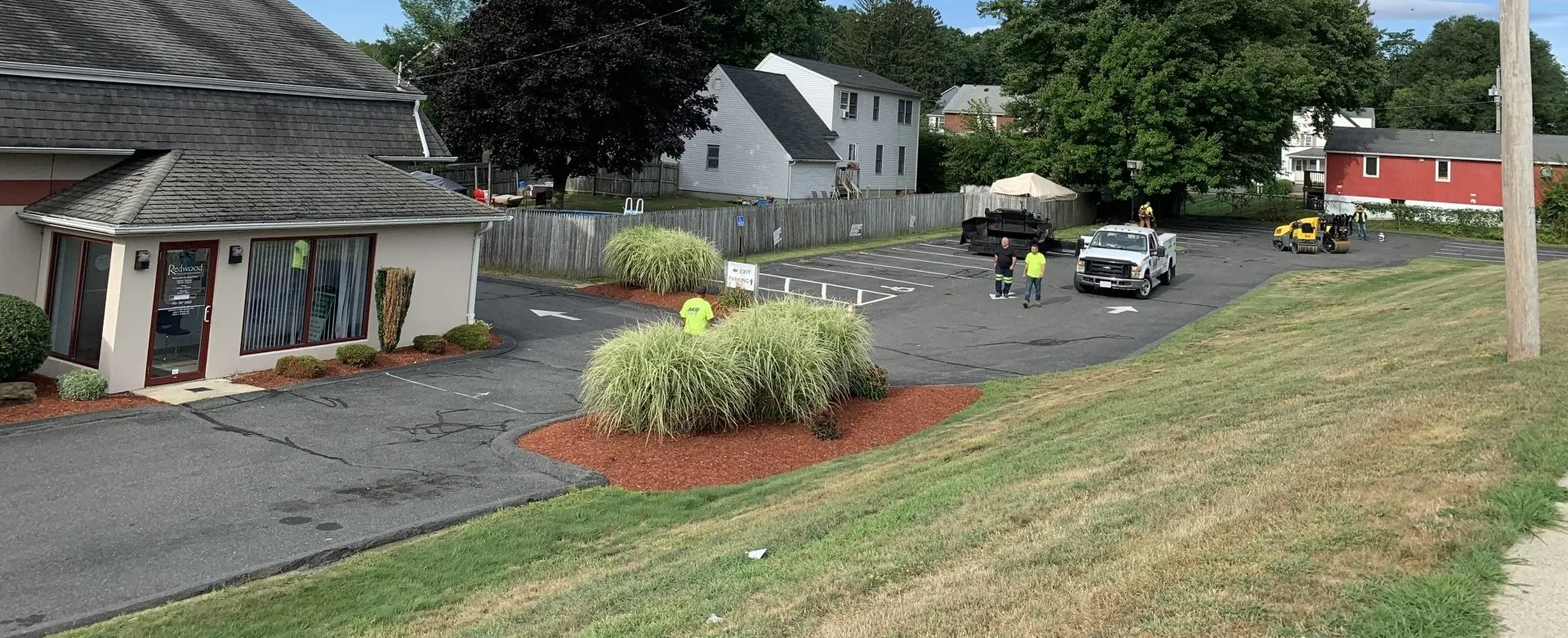 Workers near vehicles on a paved area with buildings, trees, and cloudy sky.