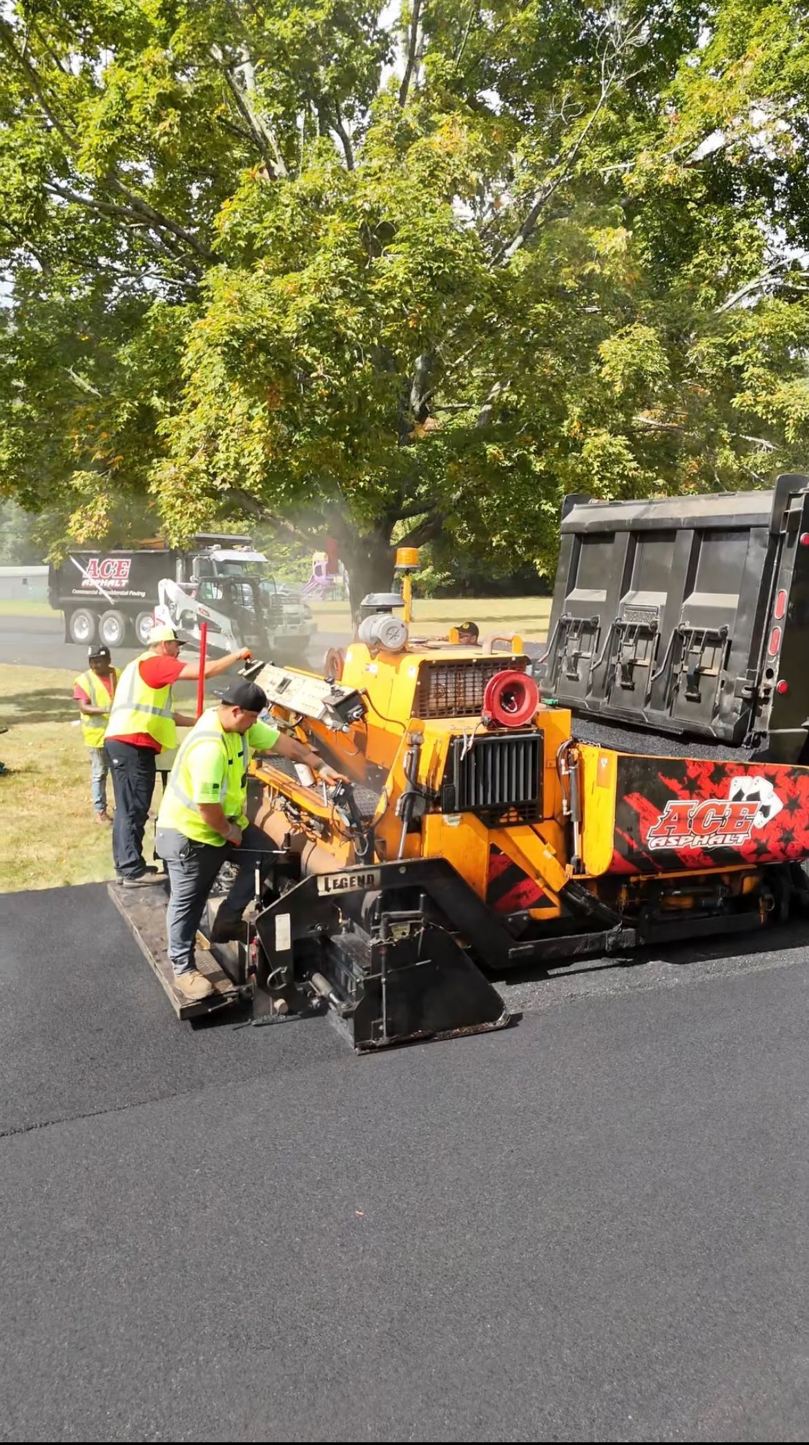 Workers paving a road with asphalt, using a large yellow paver and dump truck. Trees in background.