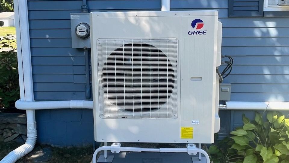 Gree heat pump unit mounted on the exterior wall of a house. It has a white frame and a wire cage over the fan.