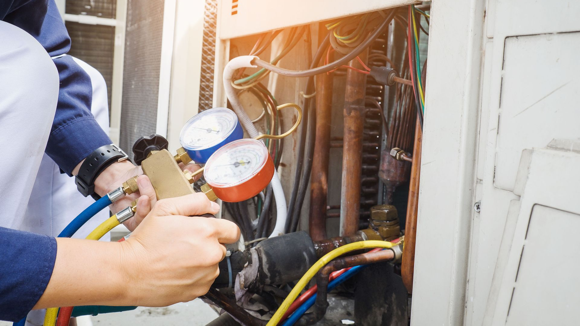 HVAC technician using gauges on an outdoor unit; blue and red hoses, wires, and metal components are visible.