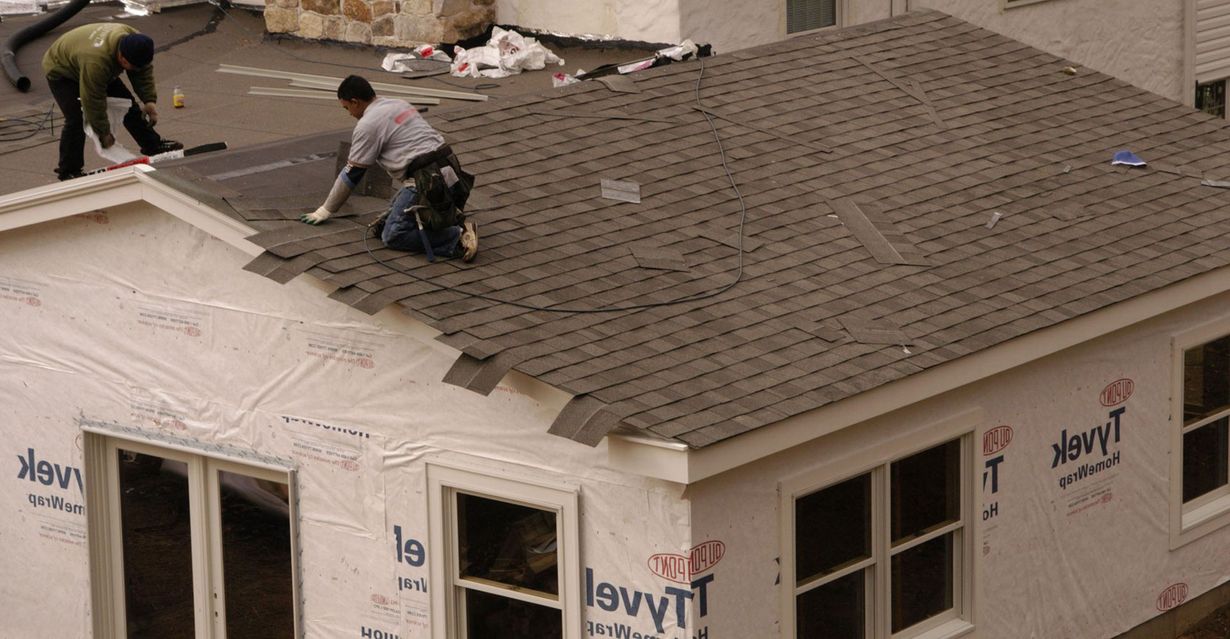 Workers installing shingles on a house roof. Building is in construction, with Tyvek wrap and windows.