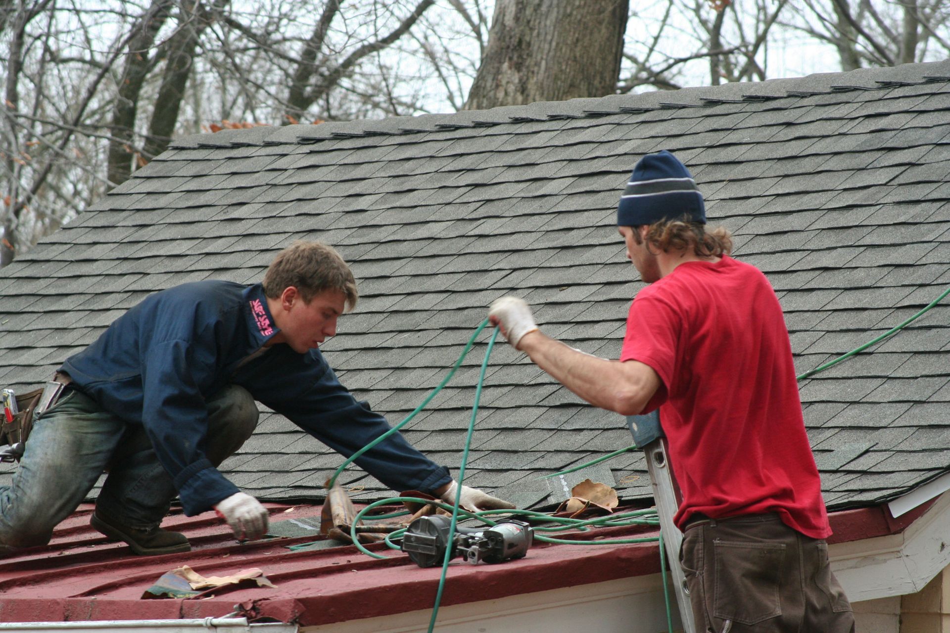 Two people on a roof working, one on a ladder, using tools on a dark roof with green rope, cloudy sky.