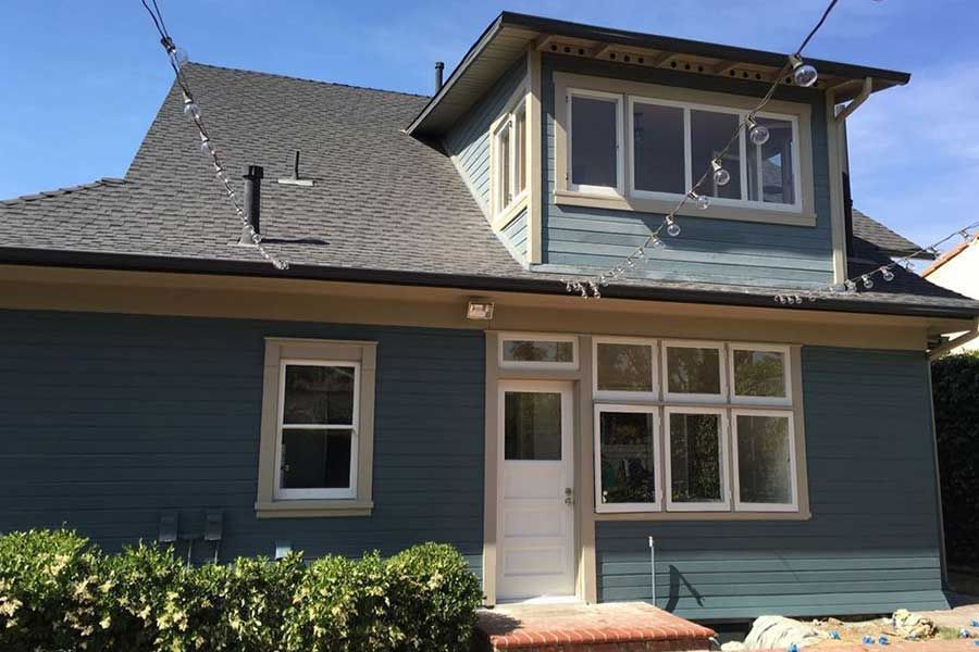 Blue house with a dormer and white-framed windows, surrounded by bushes.