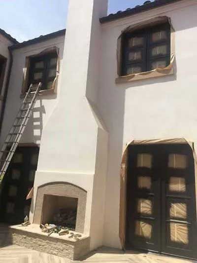 Exterior of a white stucco building with black windows and a fireplace. A ladder leans against the wall.