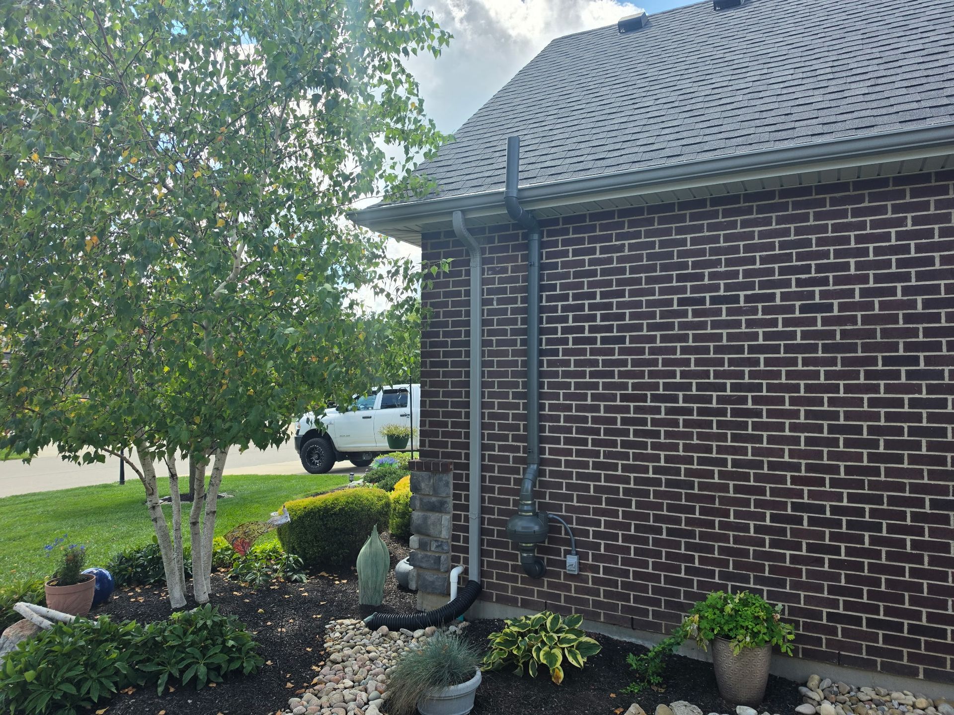 Brick house exterior with a tree, shrubs, potted plants, and a white truck in the driveway