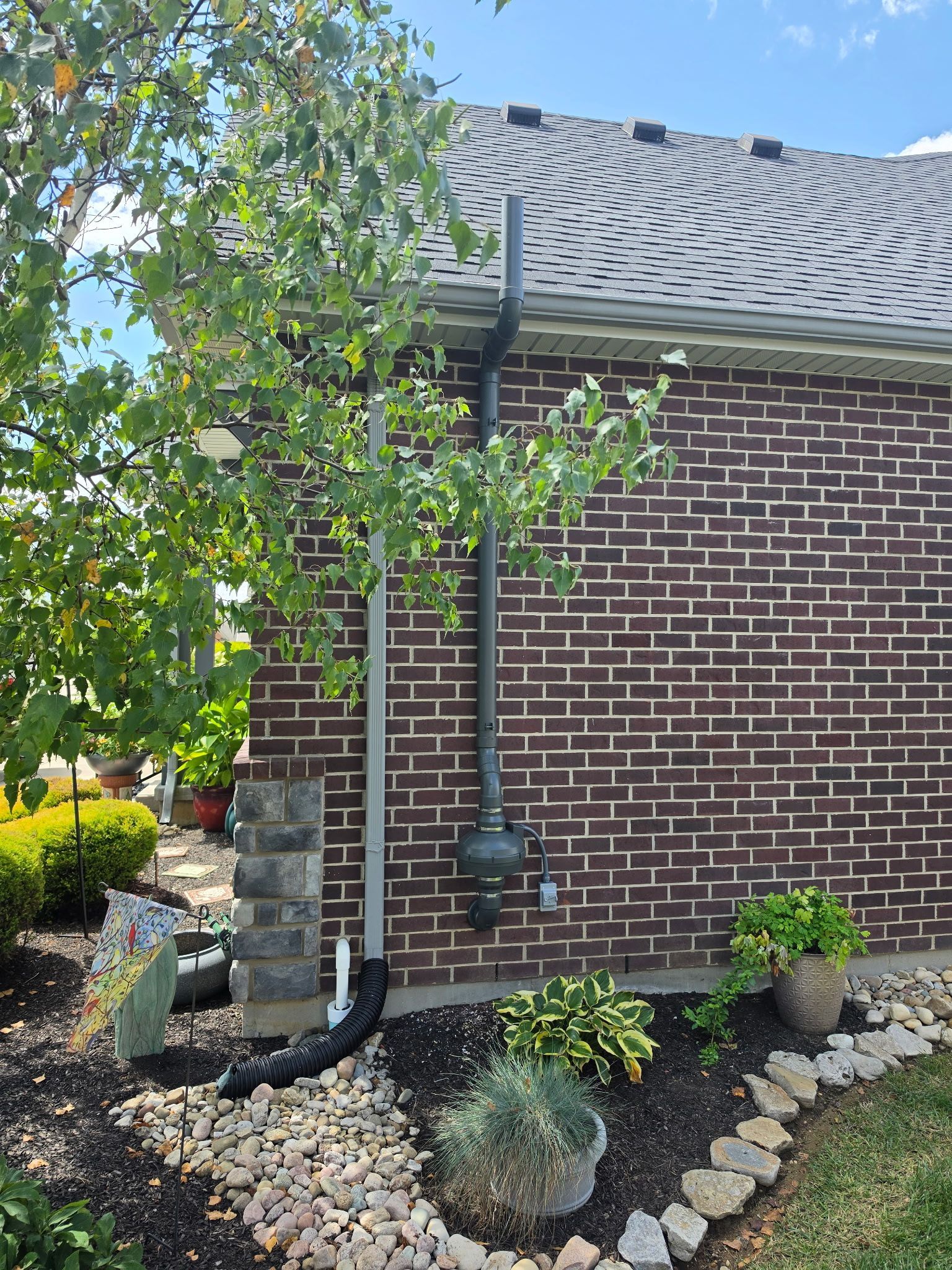 Brick house side with leafy tree, downspout, and mulch beds with shrubs and rocks