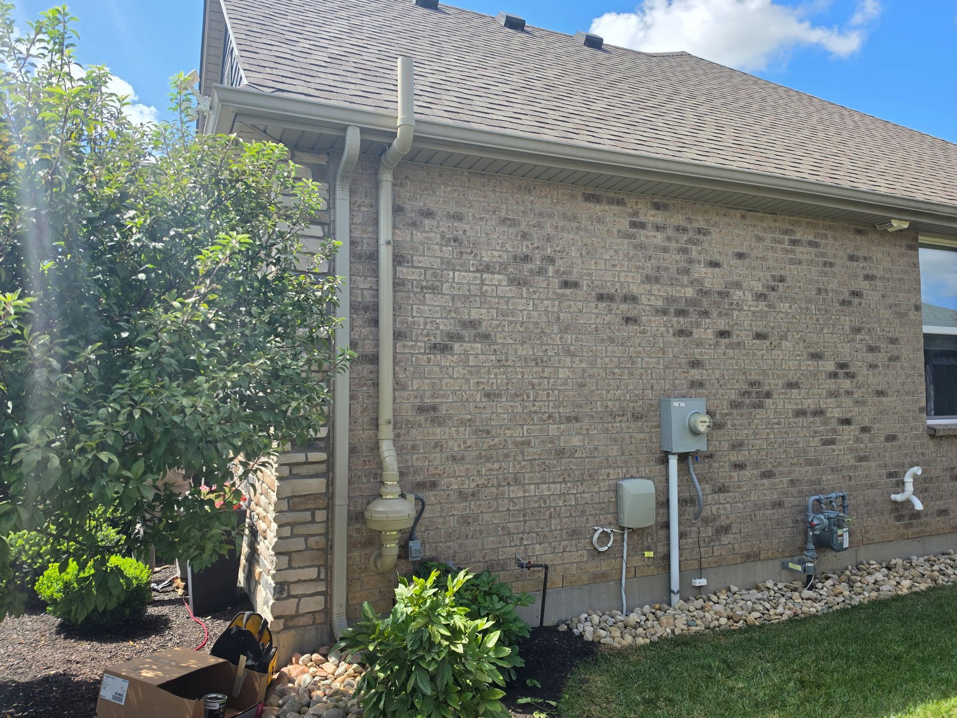 Side of a brick house with gray roof, utility meters, and a leafy tree in the yard.