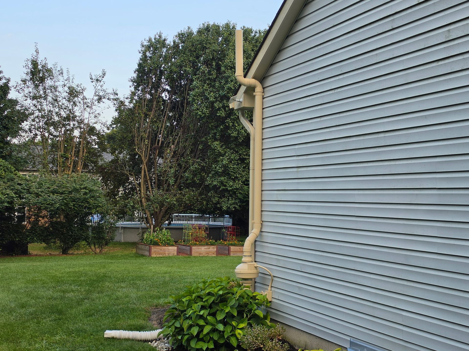 Gray-sided house corner beside green lawn and trees, with yellow downspout and brick planter.