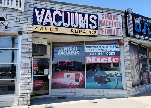Storefront with vacuum and sewing machine signs, including Miele and Central Vacuums, on a city street.