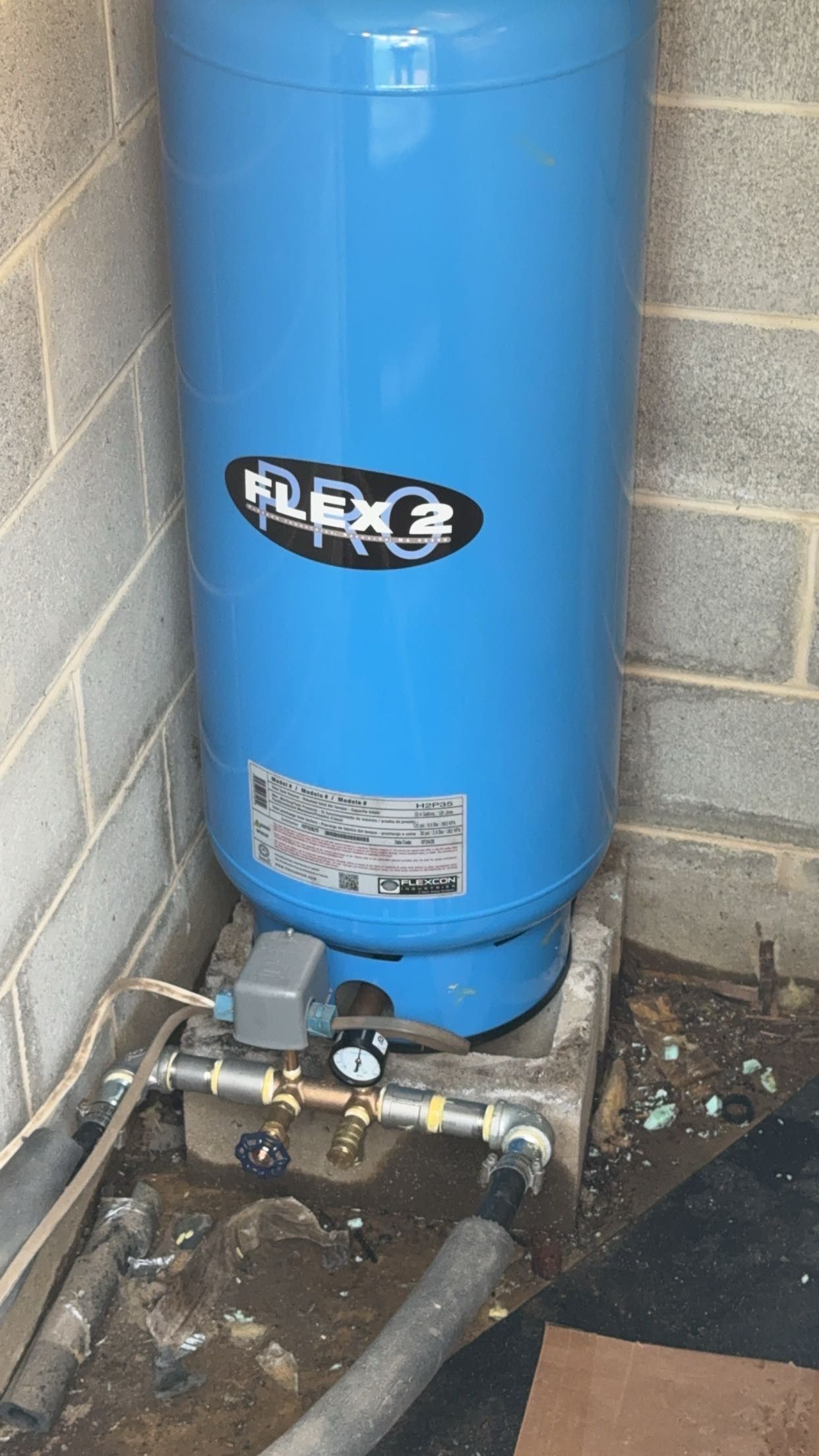Blue vertical water tank with plumbing and valves in a utility area beside a concrete wall