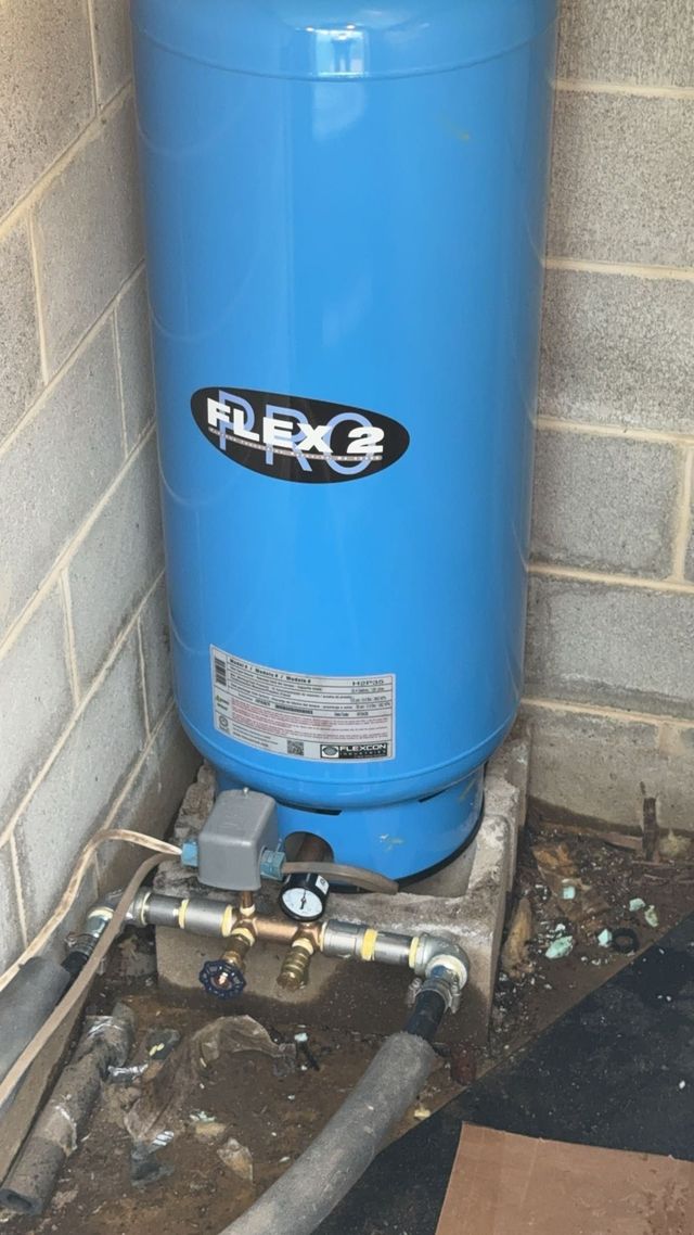 Blue vertical water tank with plumbing and valves in a utility area beside a concrete wall