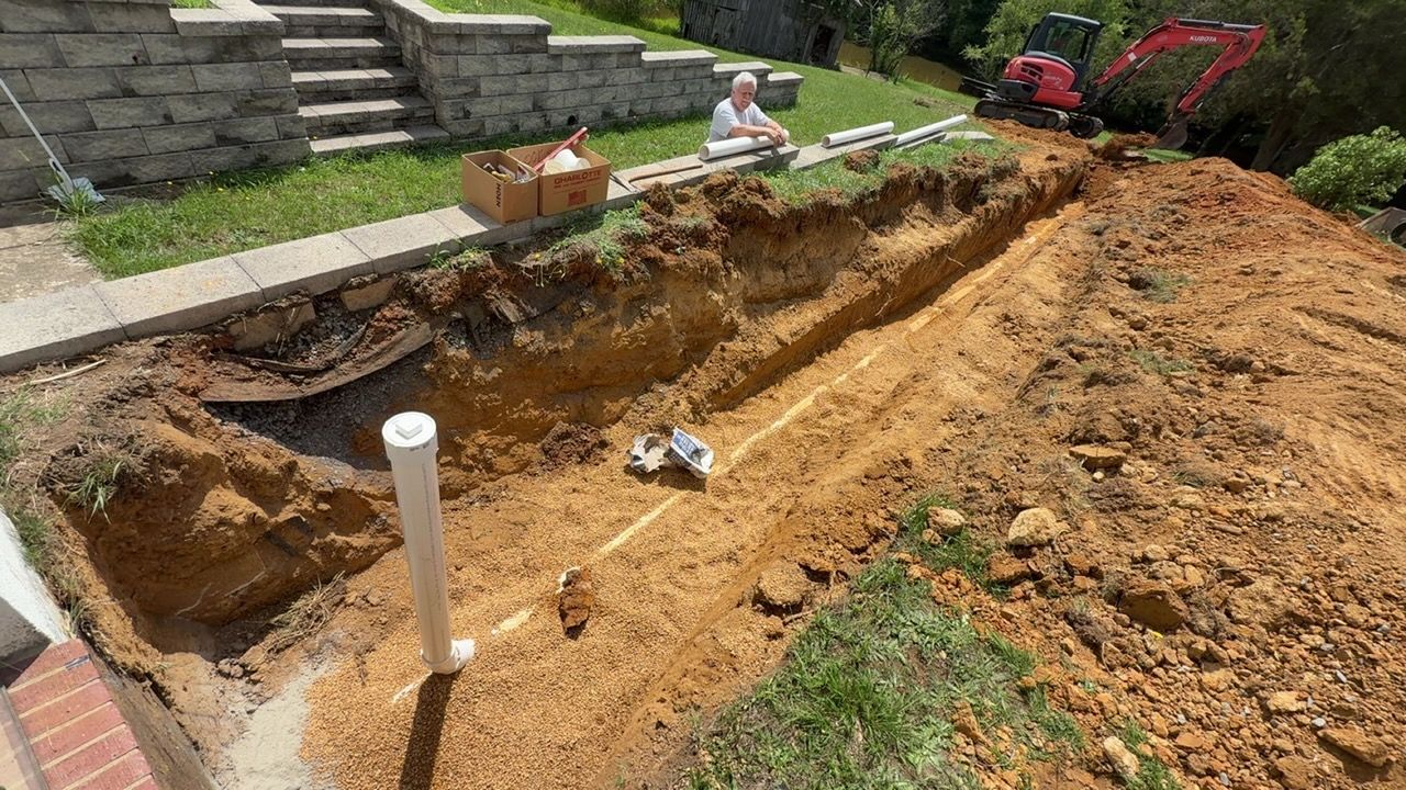 Backyard excavation beside a stone patio, with a long dirt trench and a small pipe in the foreground