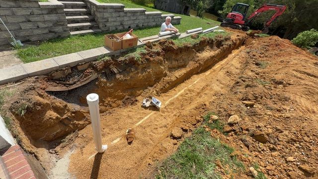 Backyard excavation beside a stone patio, with a long dirt trench and a small pipe in the foreground