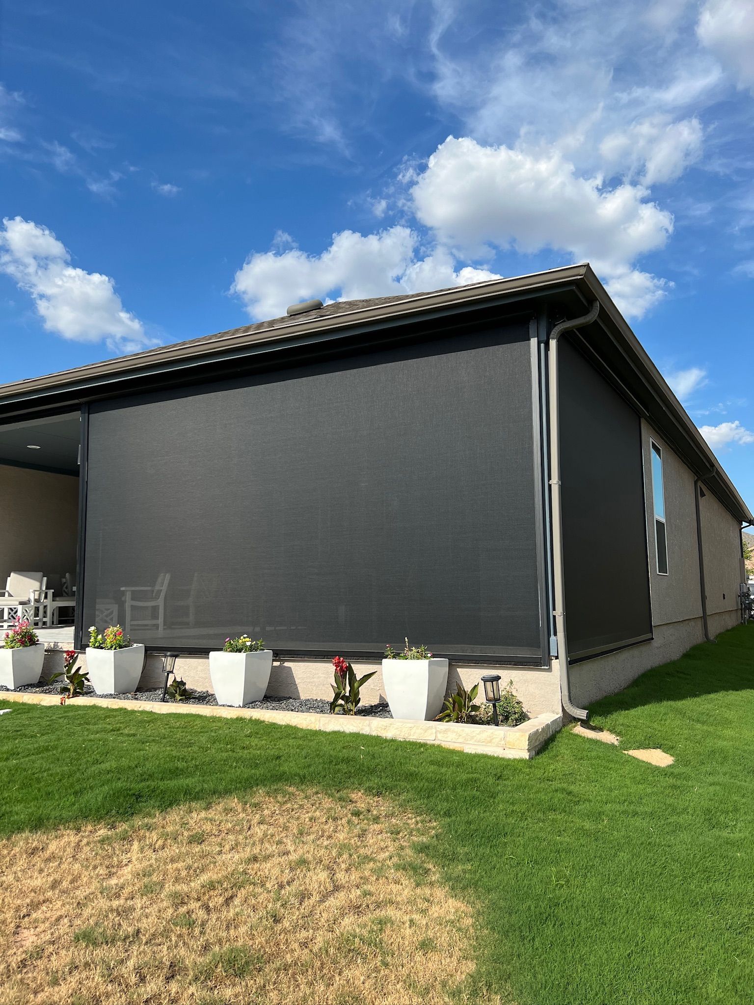 Black outdoor sunshade covering a patio, flowers in white planters, green grass, blue sky.