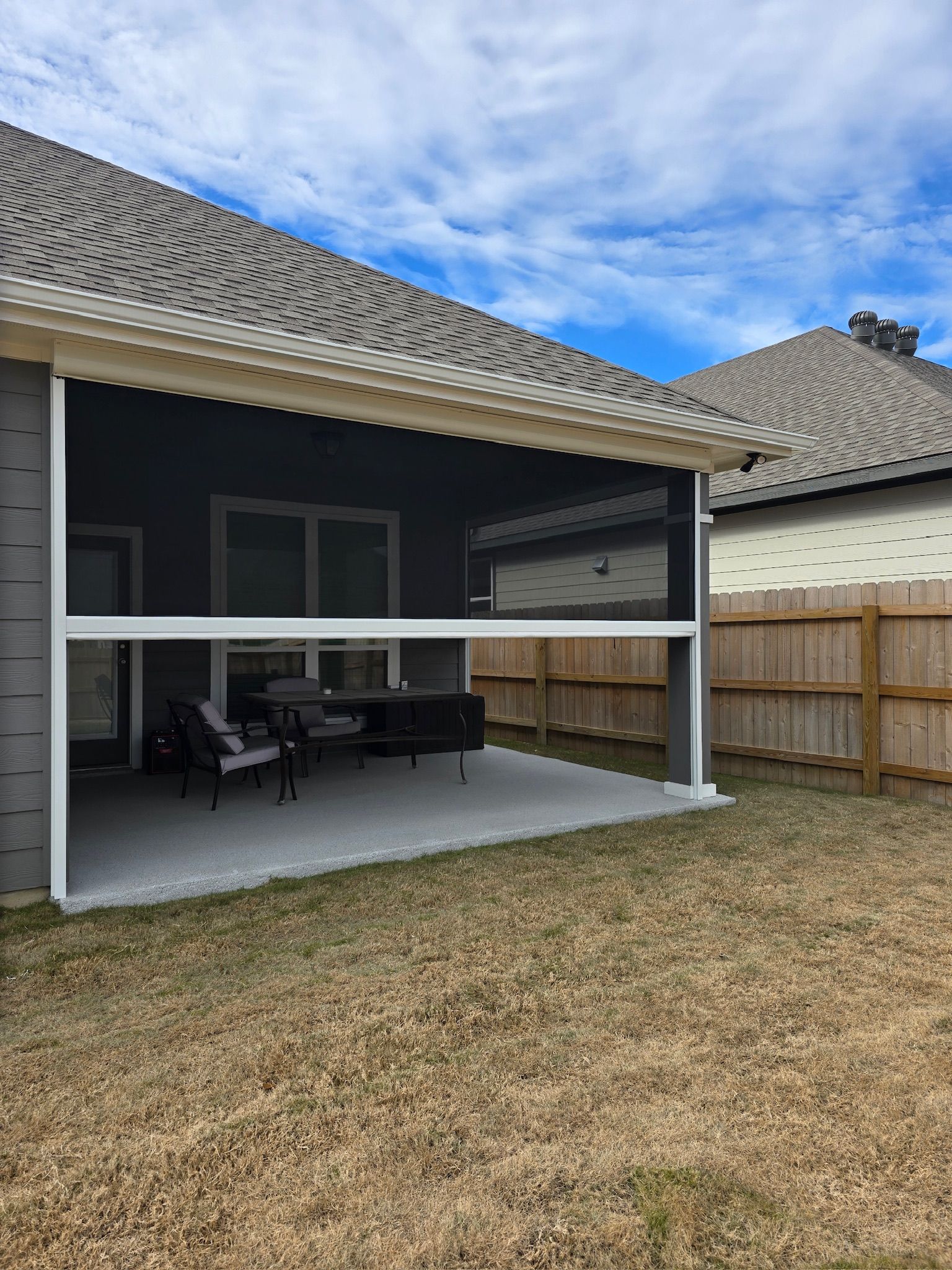 Screened-in patio with retractable shades; outdoor furniture visible. Gray walls, beige roof and fence; blue sky.