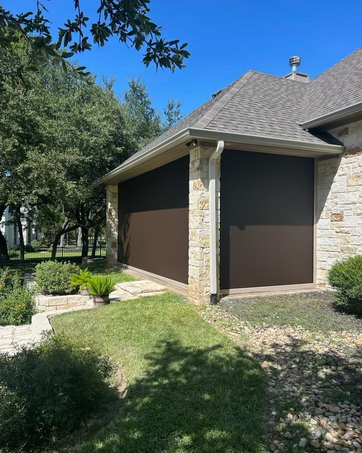 Brown retractable sun shades on a house porch, surrounded by green grass and bushes, sunny day.
