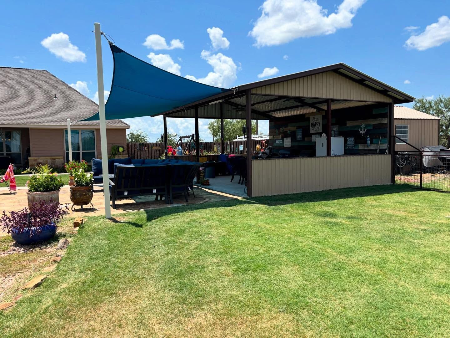 Backyard patio with shade sail, covered bar area, and green lawn under a blue sky.