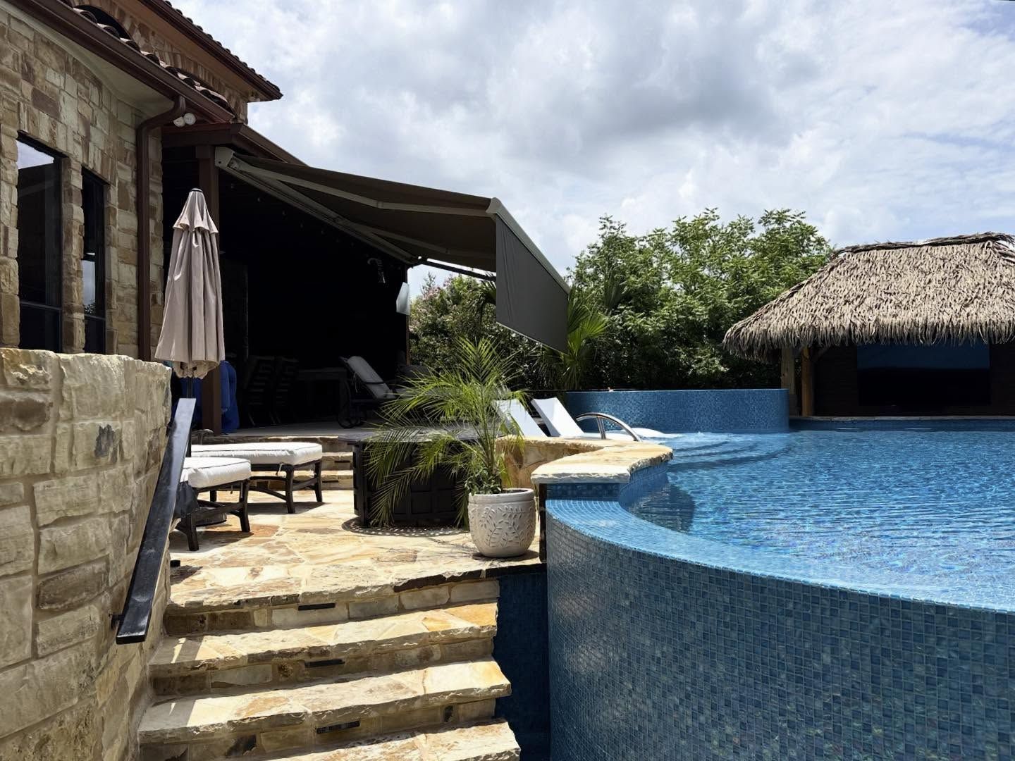 Stone steps lead to a pool with a thatched-roof swim-up bar, next to a home with an awning.