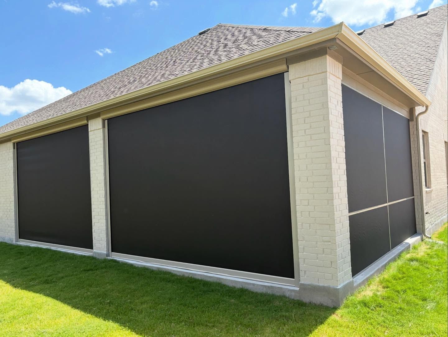 Dark shade screens cover the openings of a brick patio, against a blue sky, and green grass.