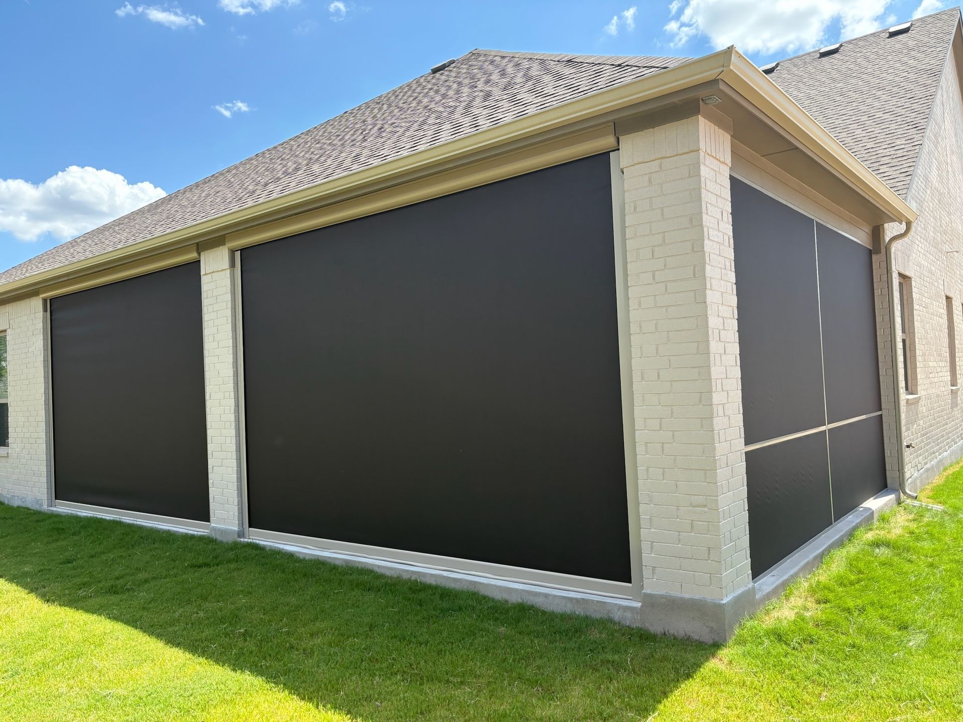 Black retractable screens on a brick patio, attached to a tan-colored house. Green grass in the foreground.