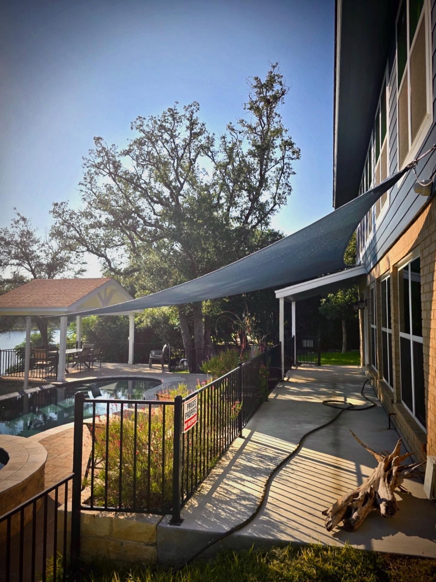 A shaded patio with a pool in the background, a blue sun sail and a dog lying on the floor.