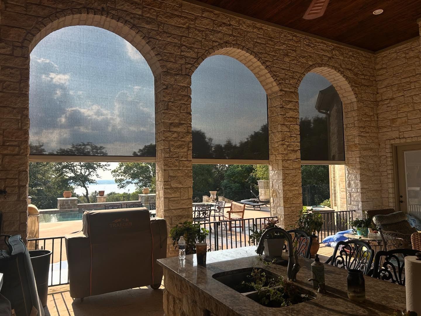 Outdoor patio with arched openings, partially shaded by black screens, overlooking a pool and lake.