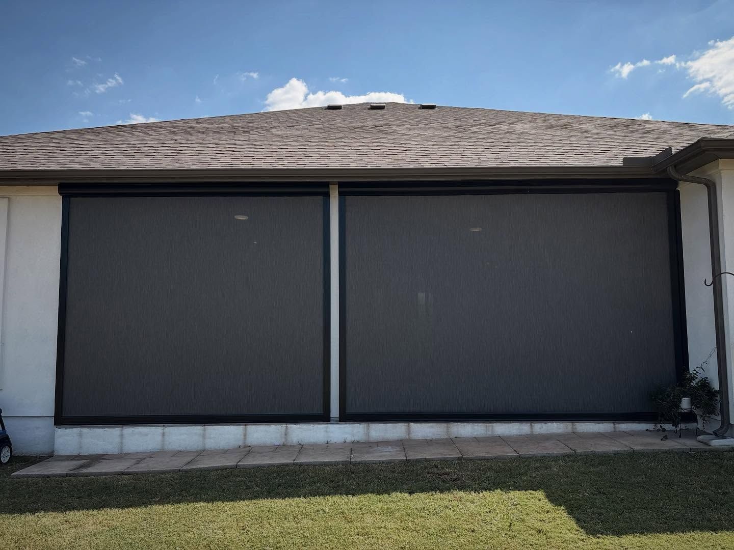 Two large, dark retractable screens on a home's exterior, shielding a patio from the sun. Blue sky.