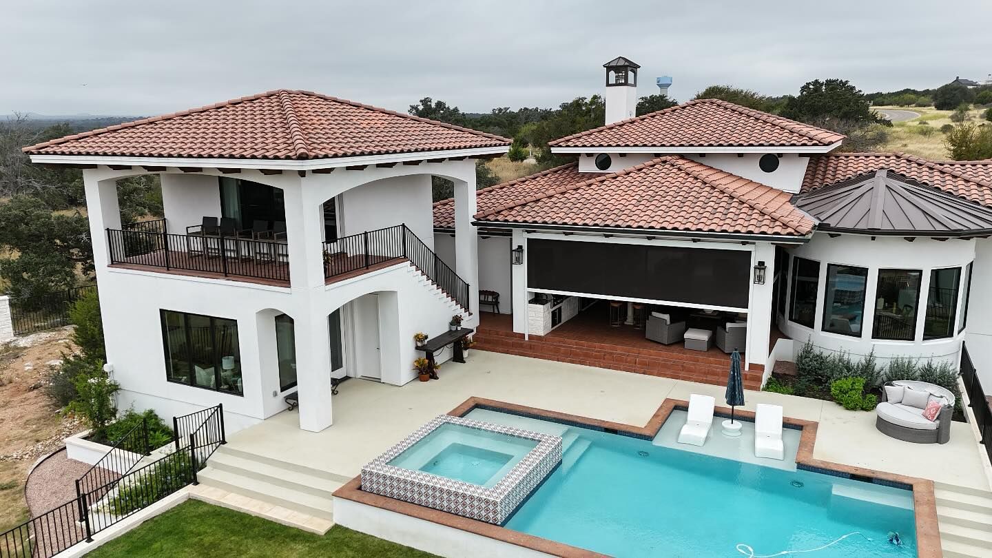 Large white house with red tile roof, pool, and patio overlooking a hilly landscape on a cloudy day.