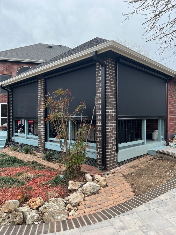 Exterior of a home with retractable black shades over a deck. Brick pillars and a brick paver pathway are visible.
