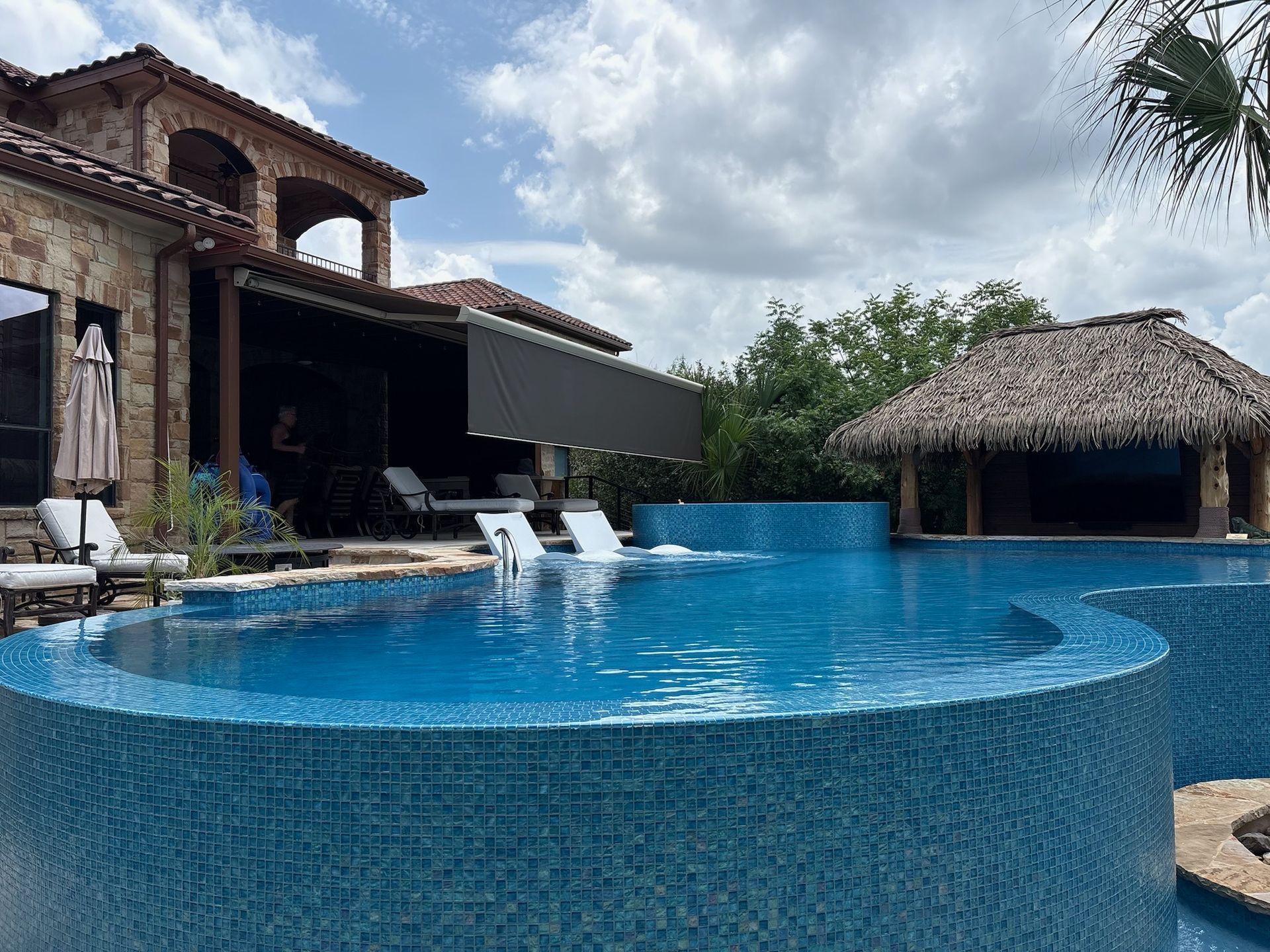 Luxury pool with turquoise tile, a thatched-roof bar, and a stone house on a partly cloudy day.