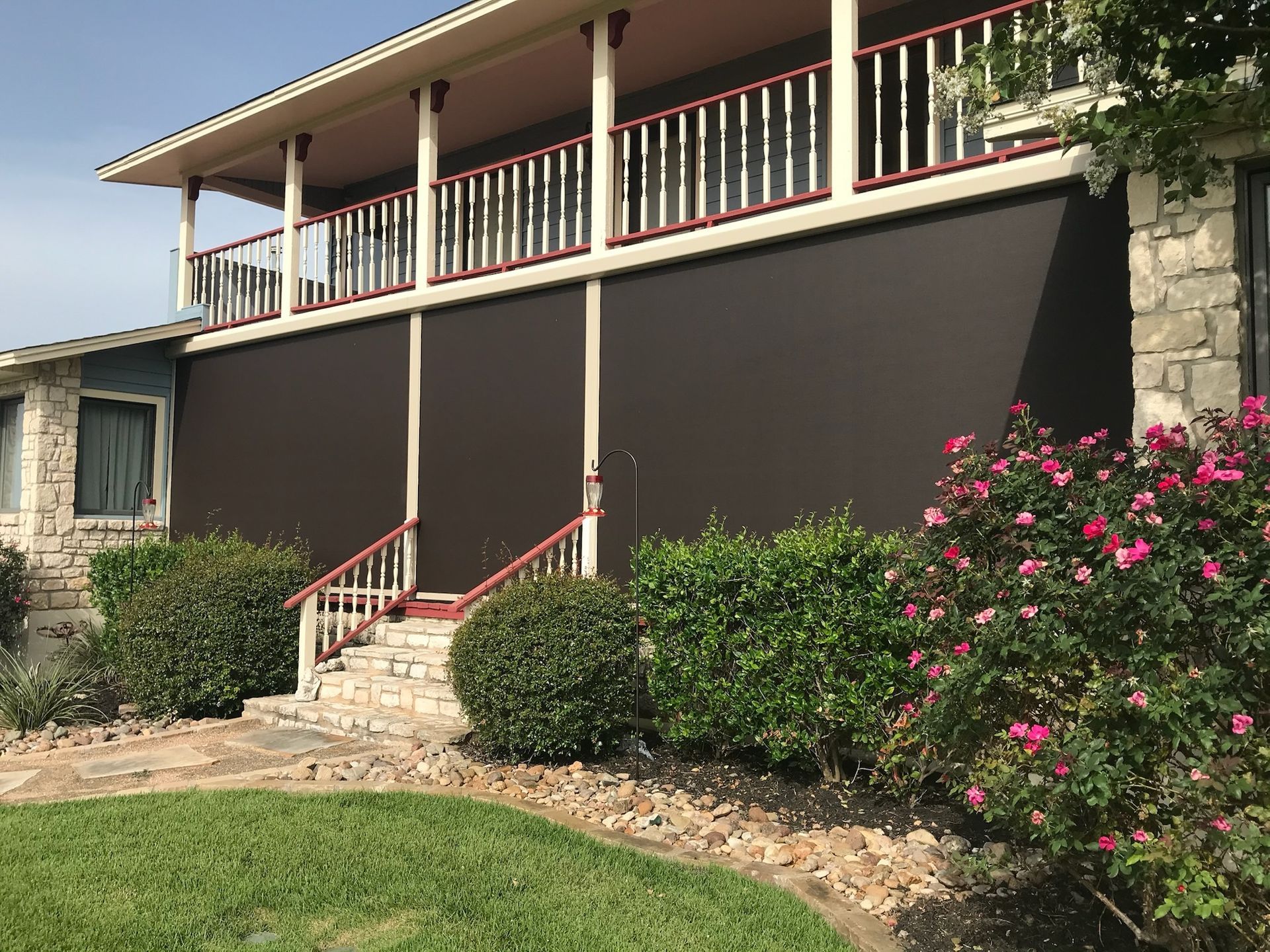 Brown sun shades cover a home's lower porch. A staircase leads up. Green bushes and grass are in the foreground.