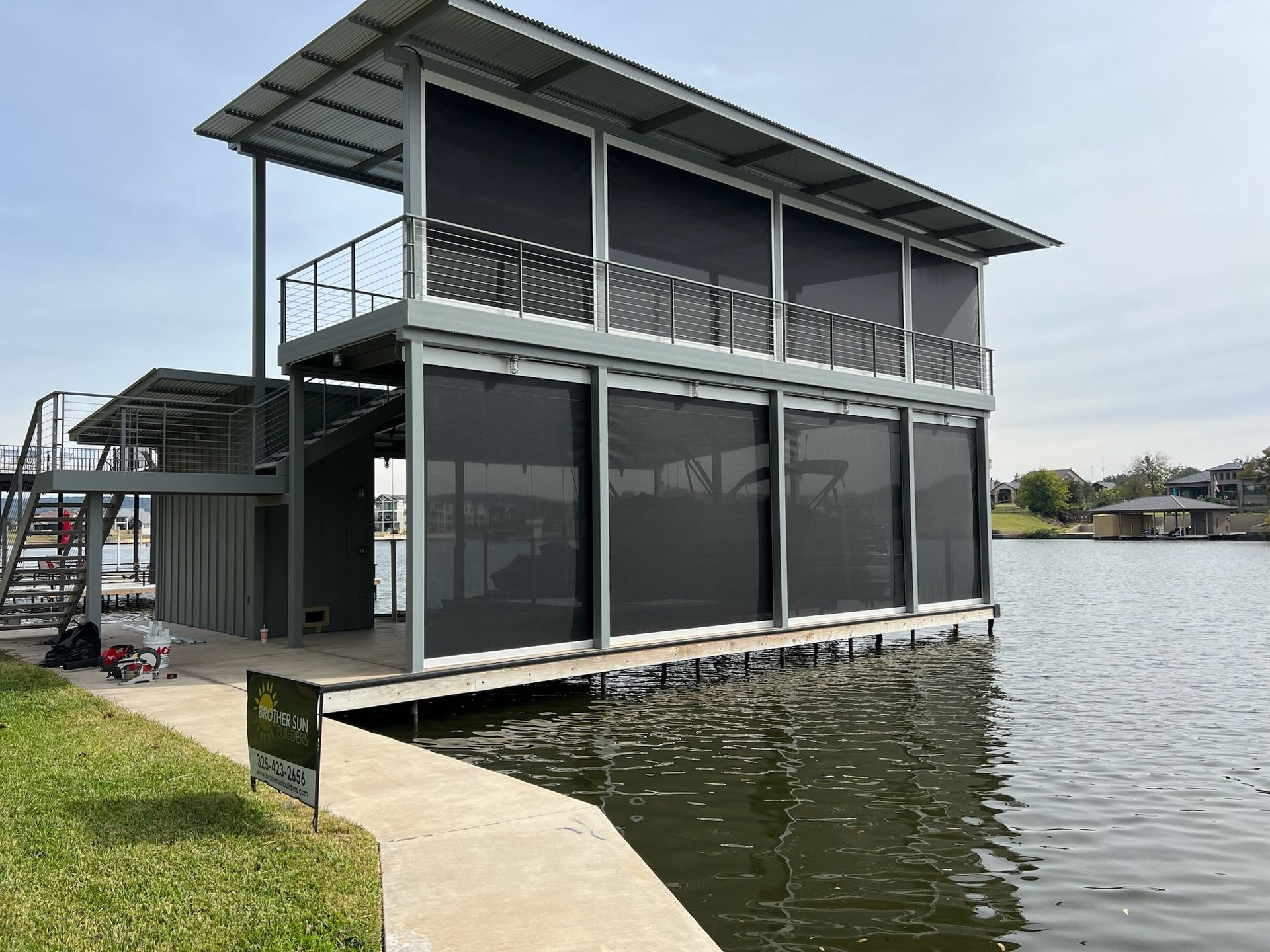 Two-story boathouse with black screens, metal railings, and a gray roof on a lake.