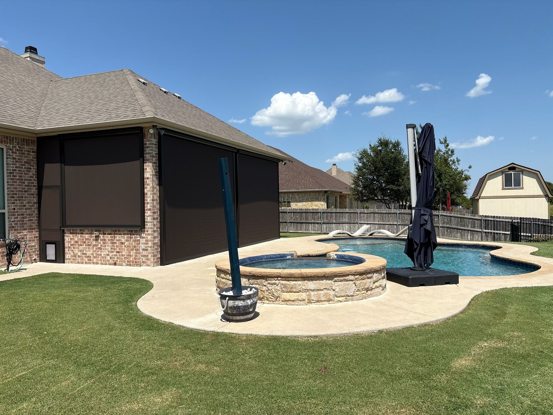 Backyard with pool, patio, and house. Black sun shades cover part of the house. Sunny day with blue sky and some clouds.