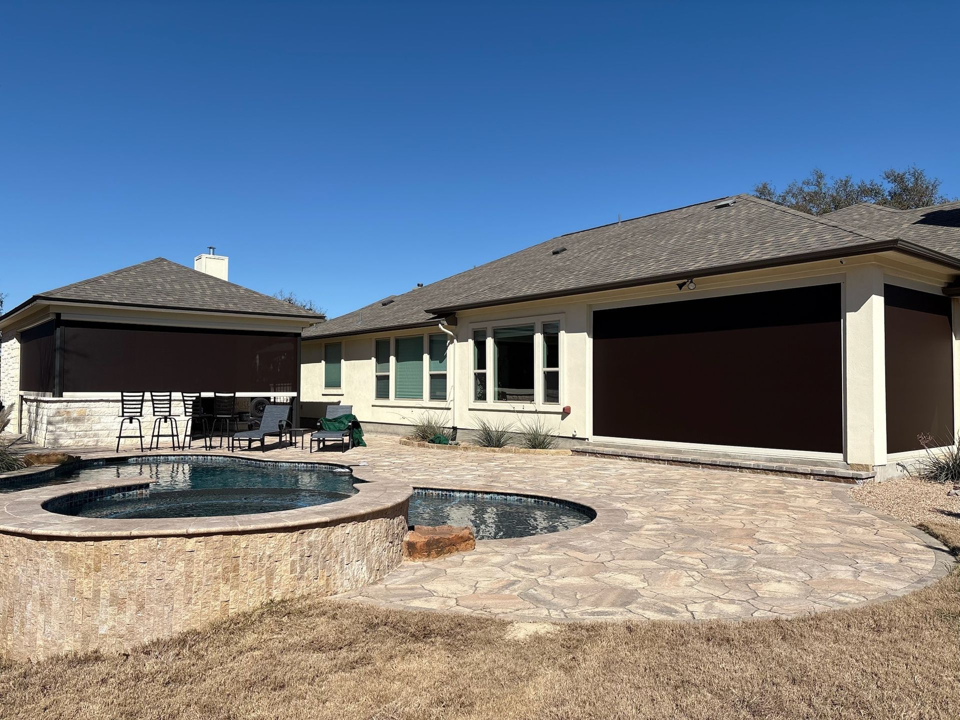 A backyard with a pool and hot tub, surrounded by a stone patio. Brown shades cover outdoor spaces.