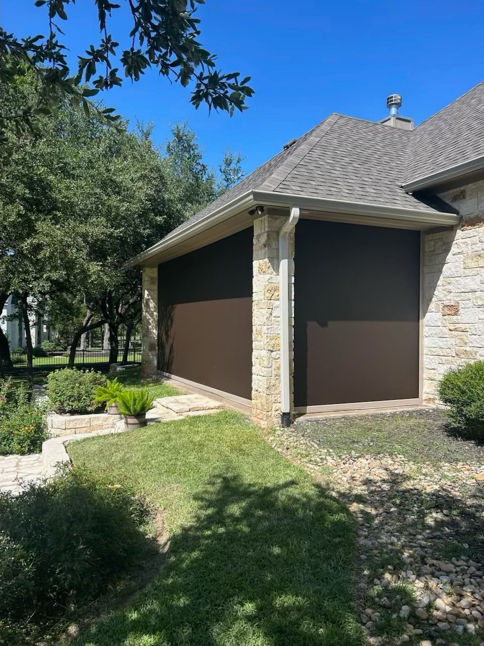 Brown garage doors on a stone and brown roof house with a green lawn and plants, under a blue sky.