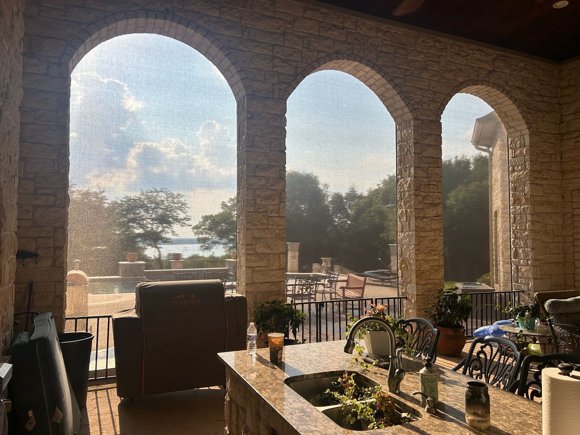 Stone archway frames a view of a lake and patio with outdoor kitchen.