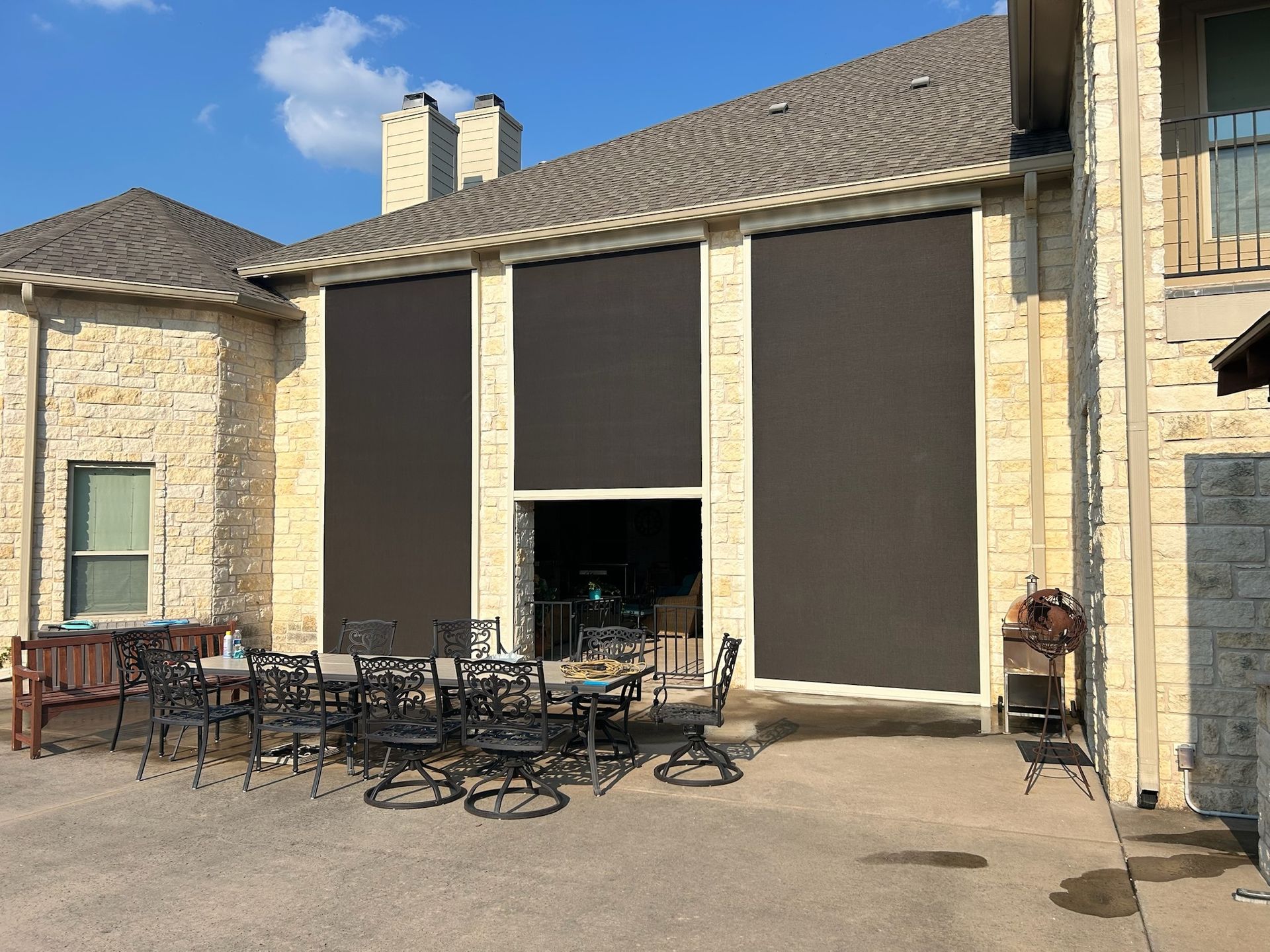 Patio with large dark-colored sun shades rolled down, covering windows. Stone house exterior, outdoor furniture.