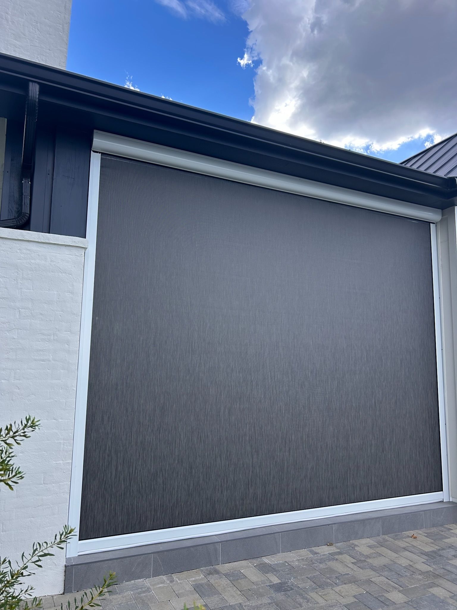 Dark gray exterior sunshade rolled down on a building with white trim, against a cloudy blue sky.